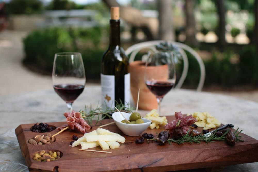 A wooden charcuterie board with cheese, meats, nuts, and olives, accompanied by two glasses of red wine and a bottle.