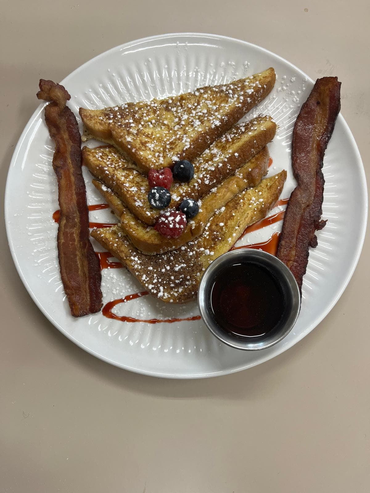 A plate of French toast triangles, bacon, fresh berries, and a small cup of syrup.