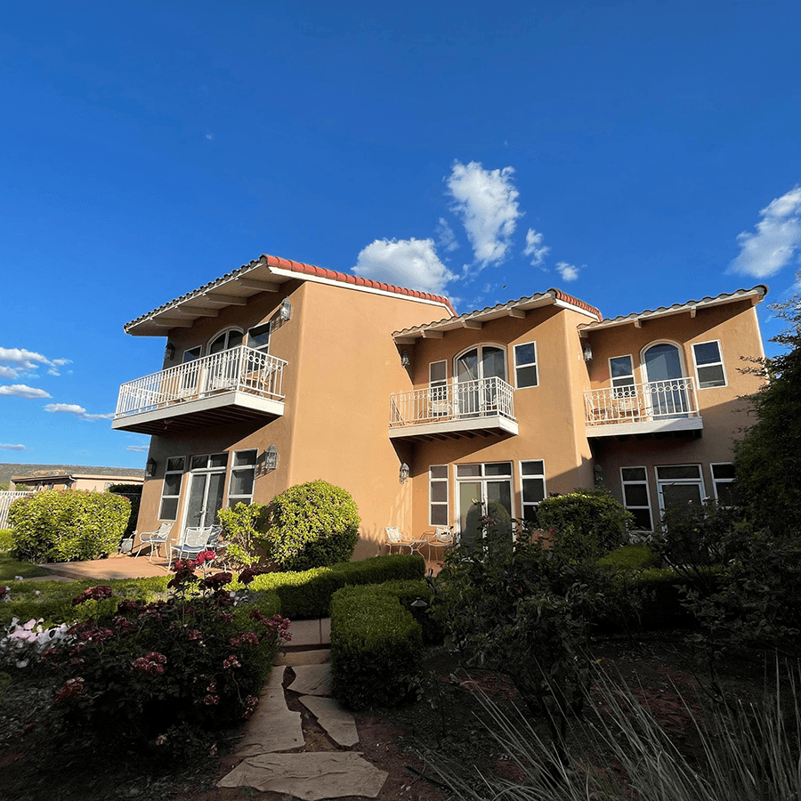 A two-story house with balconies surrounded by garden landscaping and a blue sky.