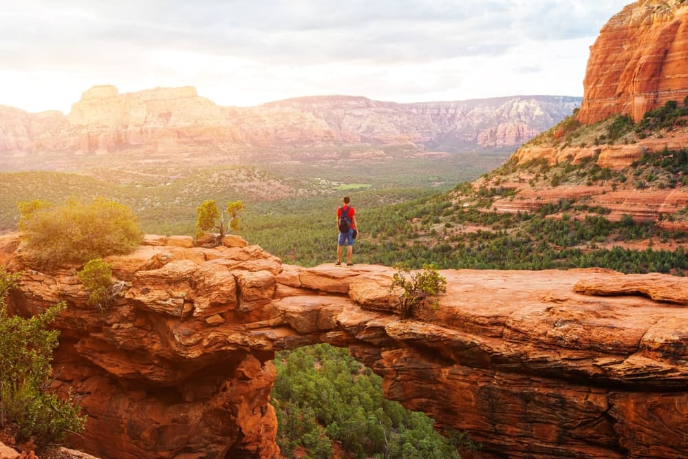 A hiker stands on a rock formation overlooking a vast canyon landscape at sunset.