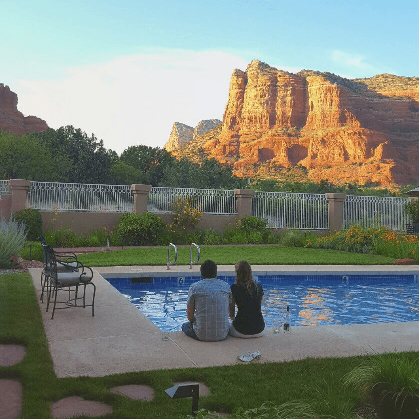 A couple sits by a swimming pool, overlooking a scenic view of red rock formations.