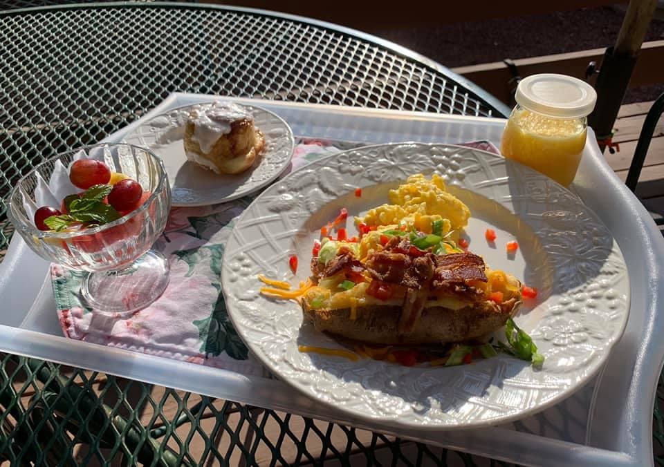 A breakfast tray featuring a loaded baked potato with scrambled eggs, bacon, and vegetables, alongside a fruit bowl and a cinnamon roll with a cup of juice.