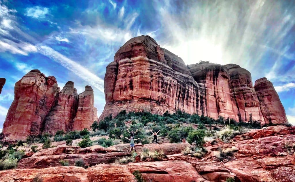 Two people stand joyfully in front of towering red rock formations under a dramatic sky.