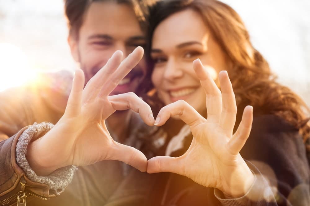 A couple smiles and makes a heart shape with their hands against a glowing background.