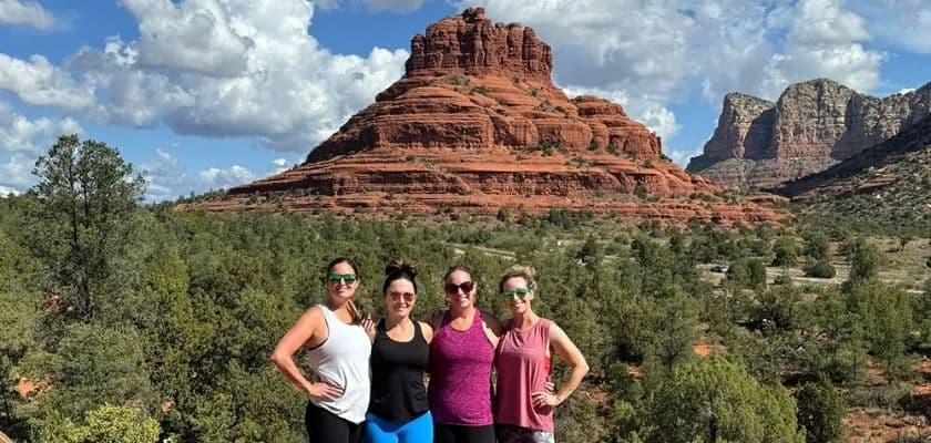 Four women pose in activewear in front of a red rock formation under a blue sky with clouds.