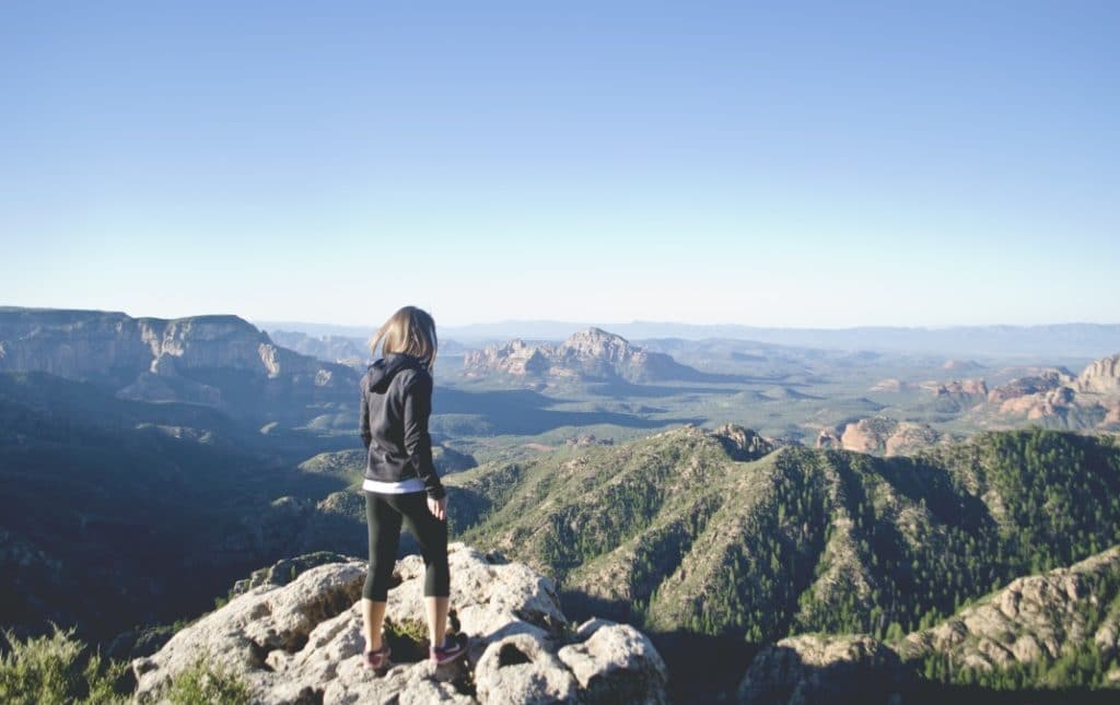 A person standing on a rocky outcrop overlooks a vast landscape of hills and mountains under a clear sky.