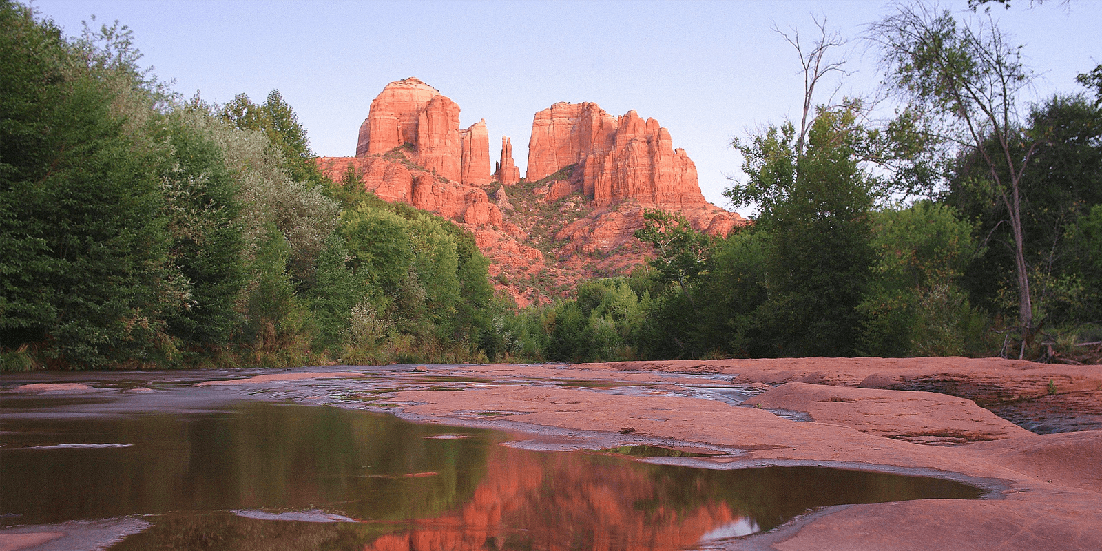 Red rock formations reflect in a calm river surrounded by lush greenery.