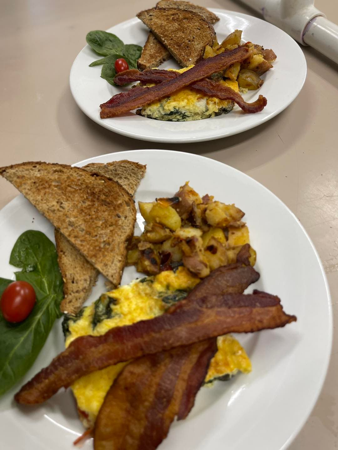 Two plates of breakfast featuring bacon, spinach omelet, toast, roasted potatoes, and cherry tomatoes.