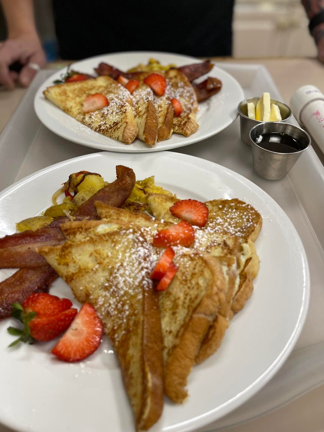 Two plates of French toast topped with strawberries and powdered sugar, accompanied by bacon and butter on the side.