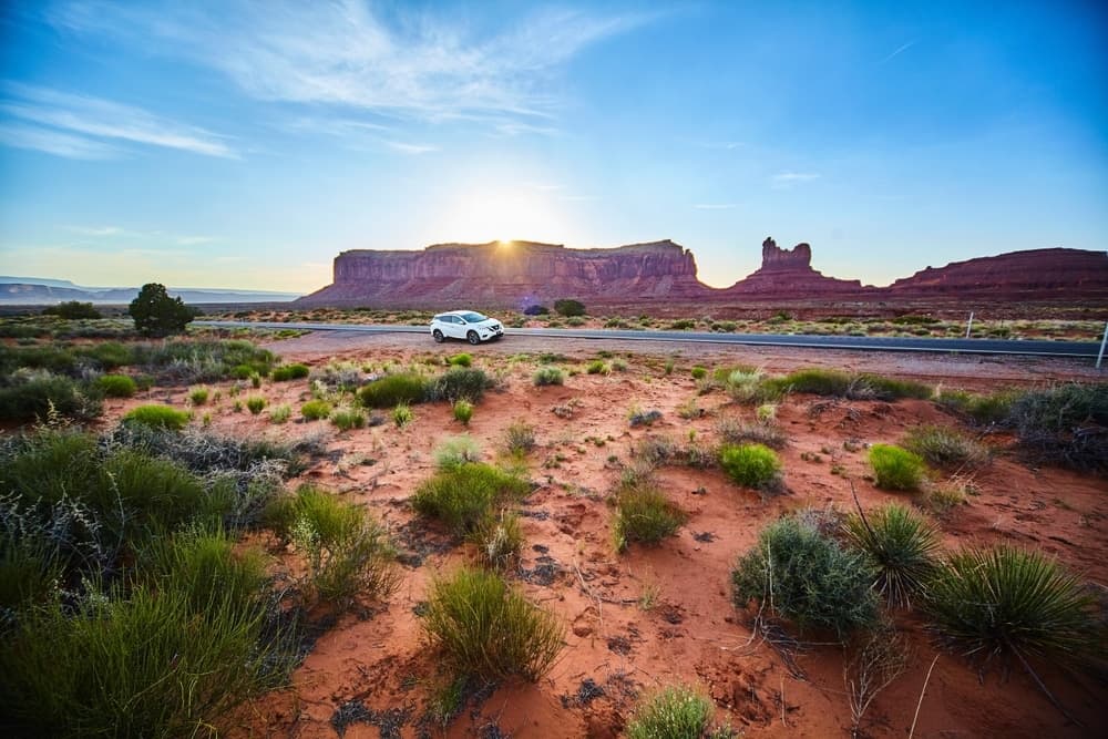 A white car drives along a road in a desert landscape with red rock formations and a sunset in the background.