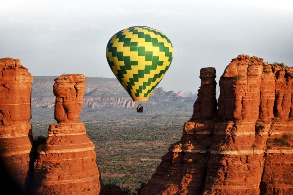 A yellow and green hot air balloon floats between red rock formations against a backdrop of green landscape.