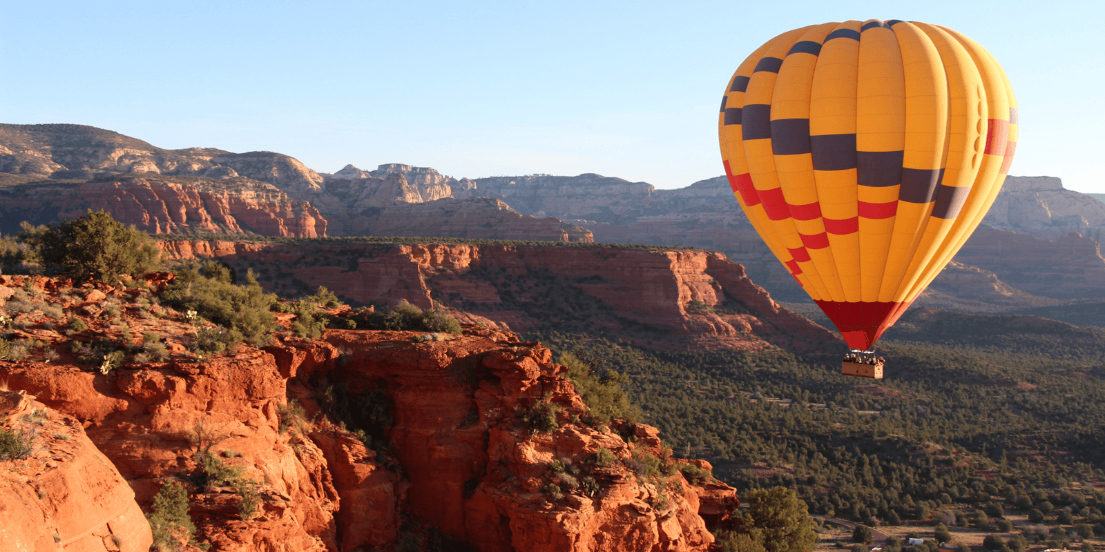 A colorful hot air balloon floats over red rock cliffs and green trees.