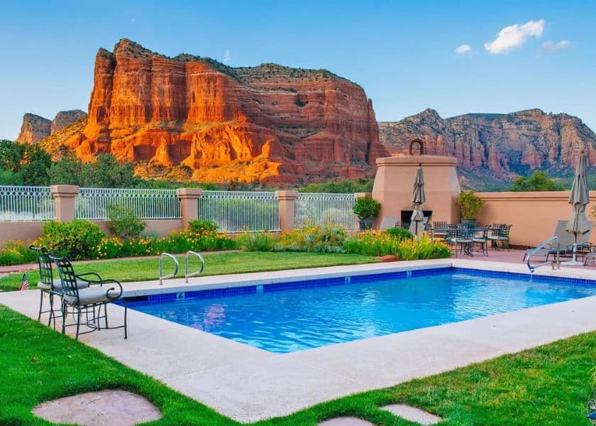 A sparkling pool with lounge chairs is surrounded by vibrant green grass and stunning red rock formations under a clear blue sky.