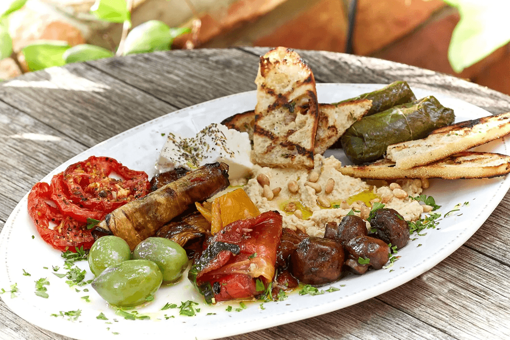 A colorful platter featuring grilled vegetables, olives, hummus, bread, and stuffed grape leaves.