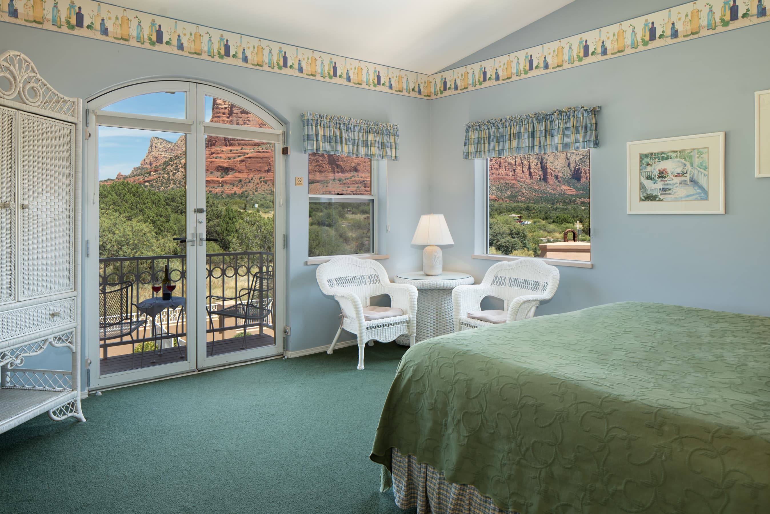 A light blue room with a green bedspread, white wicker furniture, and a balcony offering stunning views of red rock formations and lush trees.