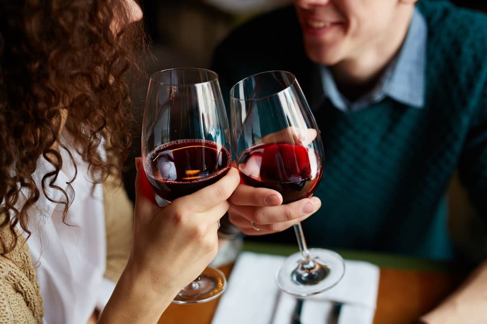A couple clinks glasses filled with red wine in a cozy restaurant setting.