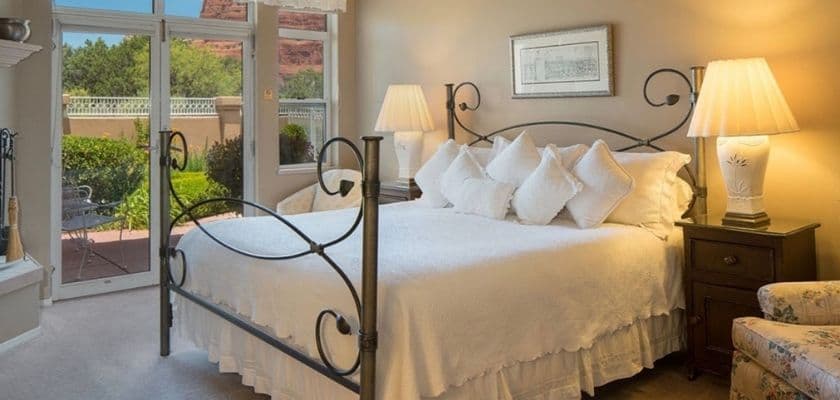 a bright cozy bedroom with a wrought iron bed, white bedding, soft lamps, and a view of bushes and red rock landscape through large windows