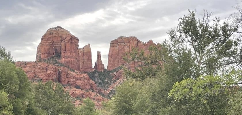 Cathedral Rock formations in Sedona rise above green trees beneath a cloudy sky.